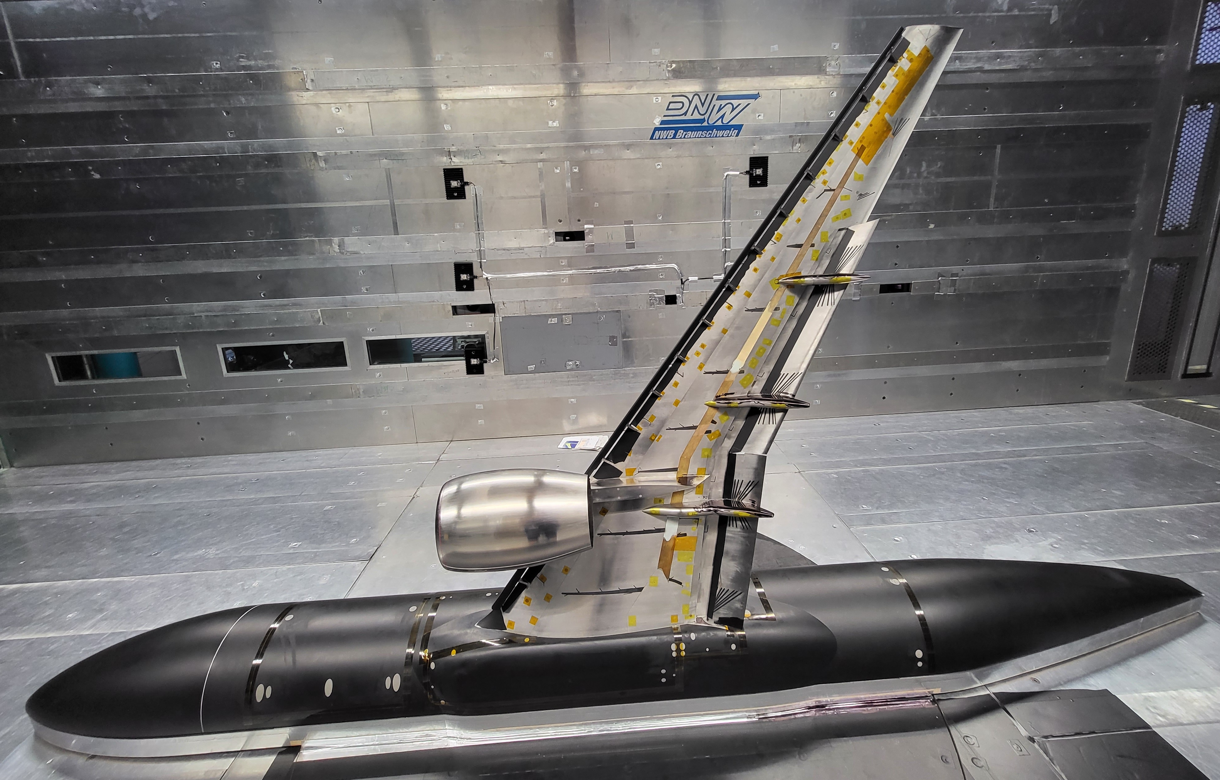 A silver-colored airplane research model sits in a wind tunnel facility. The model consists of a section of a scaled-down fuselage on the floor, wi...