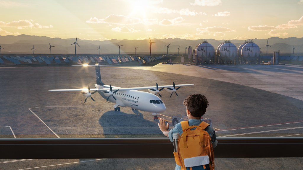 An artist’s concept that depicts an airliner with two propeller engines on each wing resting on airport tarmac. The aircraft is viewed through a window and, in the foreground, a small child with dark hair and a yellow backpack looks at it, with his hand resting on the glass. Sustainable infrastructure is visible in the far background, including wind turbines and large tanks with LH2 written on them, the formula for liquid hydrogen, a potential alternate fuel source.