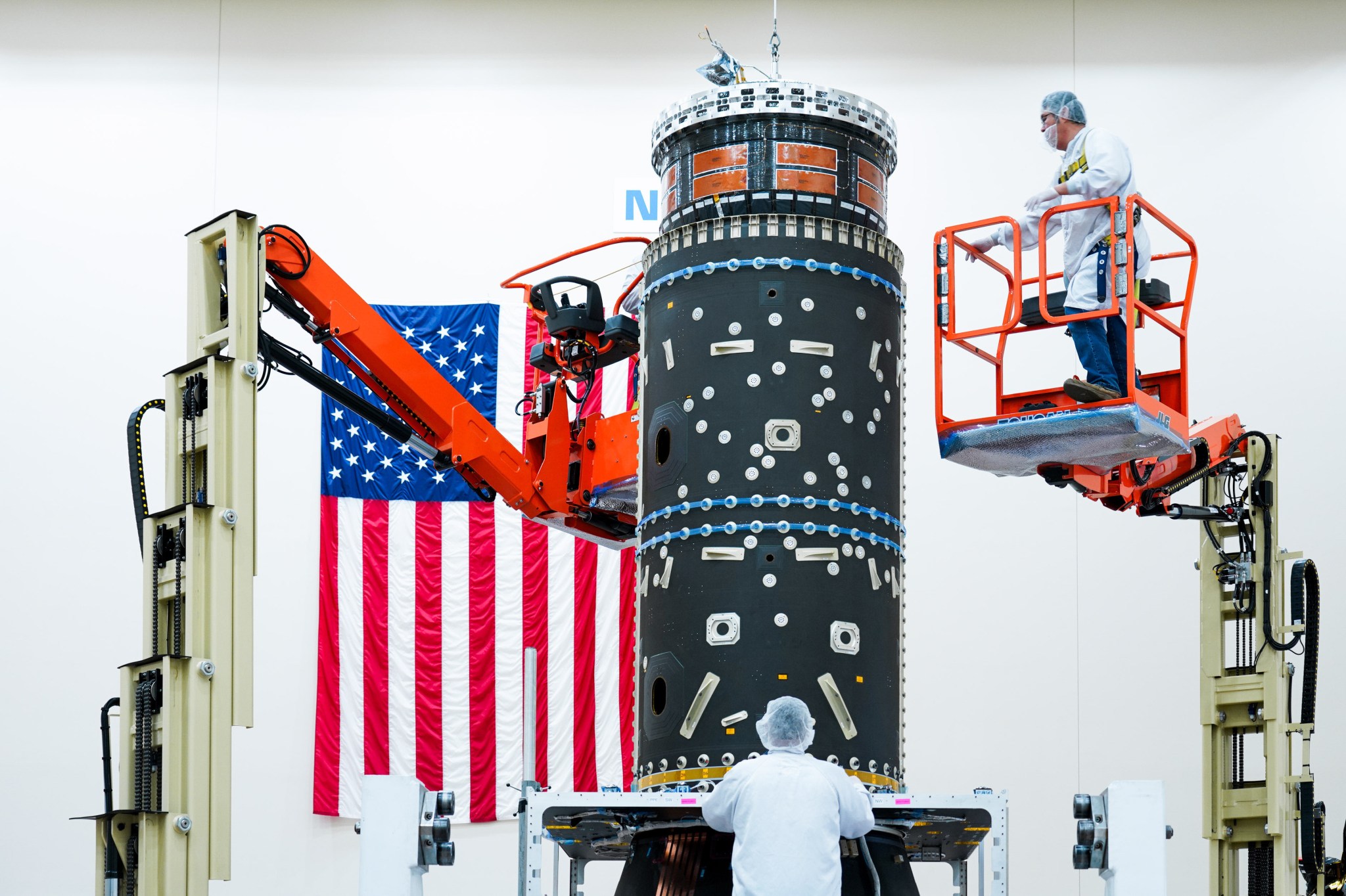 Technicians work on the installation of hardware designed to house Gateway’s xenon fuel tanks, an integral component of its advanced electric propulsion system. The setup is conducted in a cleanroom environment, with one technician on an elevated lift and another standing at the base of the structure. An American flag is displayed prominently in the background.