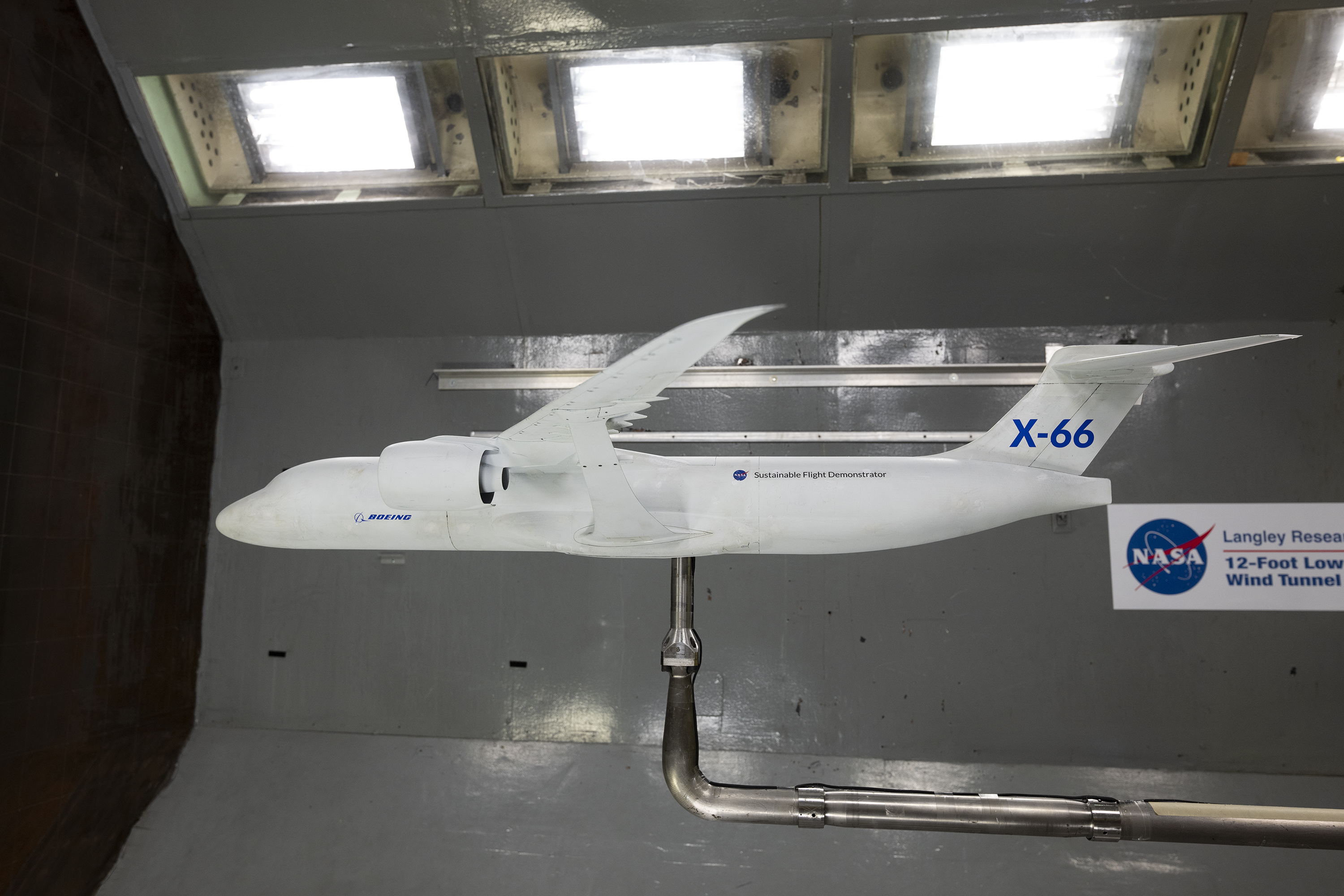 A white model of an airplane mounted on top of a “L” shaped silver pole with a background of shiny grey walls, there are bright white lights on...