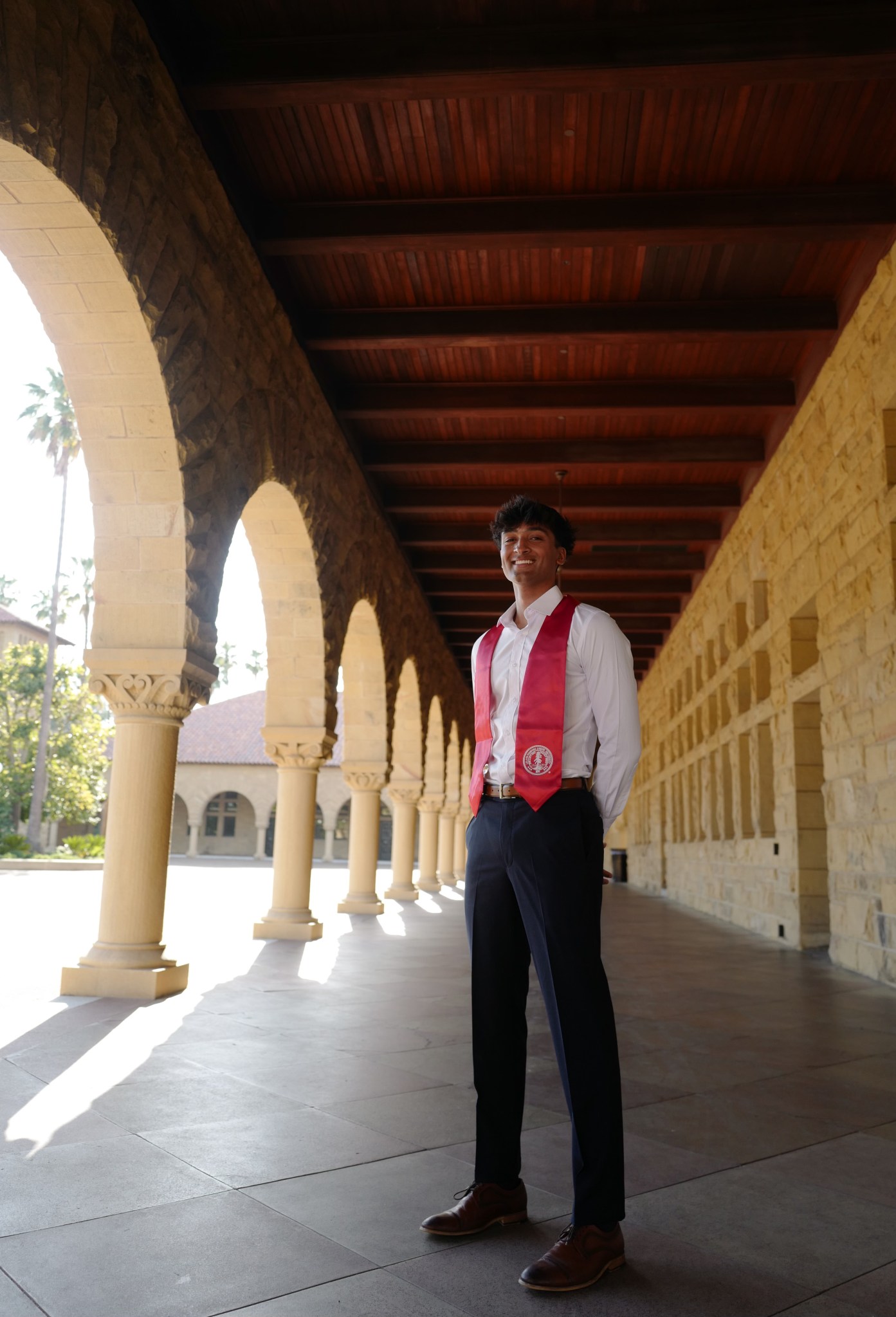 A young man standing in a shaded colonnade with stone arches, wearing a red graduation stole over a white shirt and dark trousers.