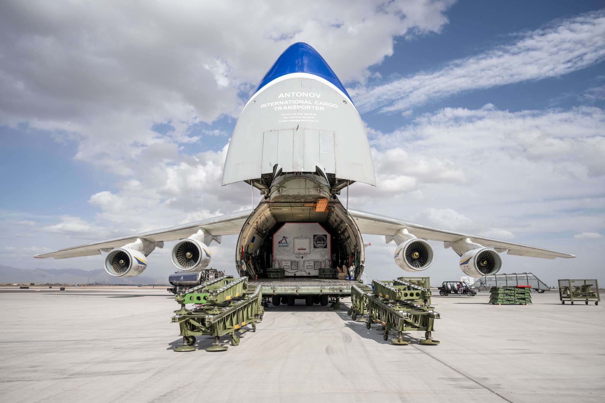 A large cargo aircraft with its nose lifted open reveals HALO's wrapped container. The container is marked with the logo for NASA's Artemis campaign. The scene is set at an airport during late afternoon.