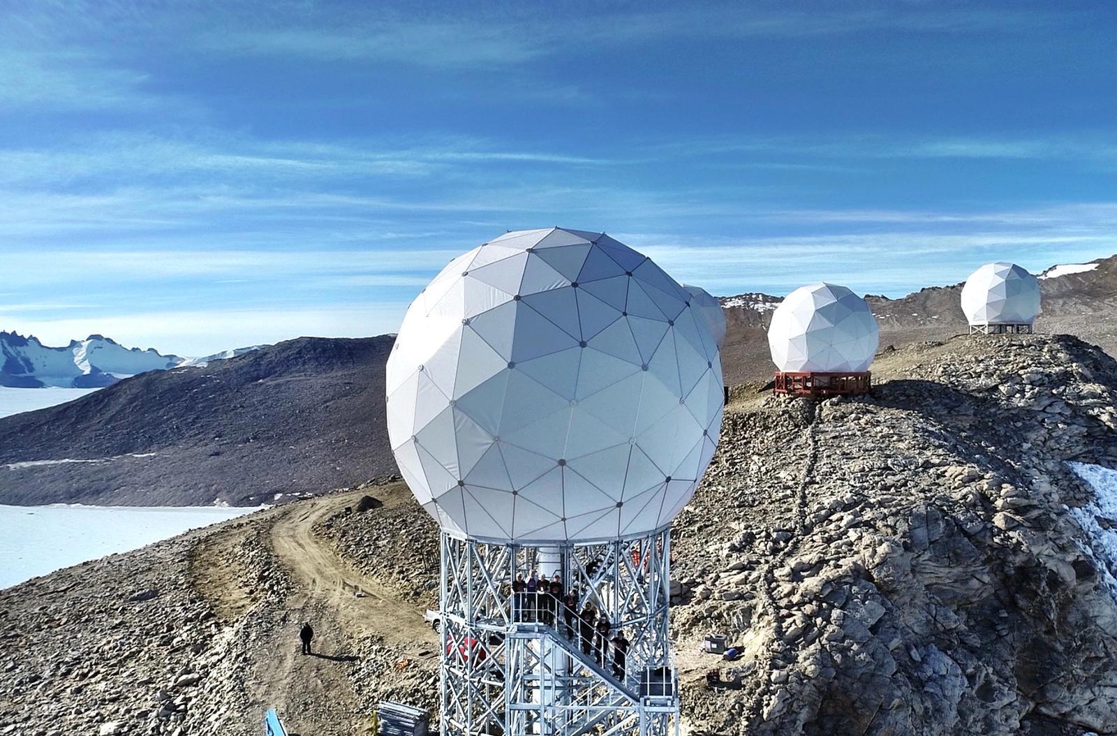 A large, white geodesic dome protecting a ground station antenna sits on a raised platform labeled KSAT at the Troll Satellite Station in Antarctica. The rocky, snow-dusted terrain sits in the foreground of an expanse of ice and a clear blue sky. A small group of people is visible at a distance near the edge of the plateau.