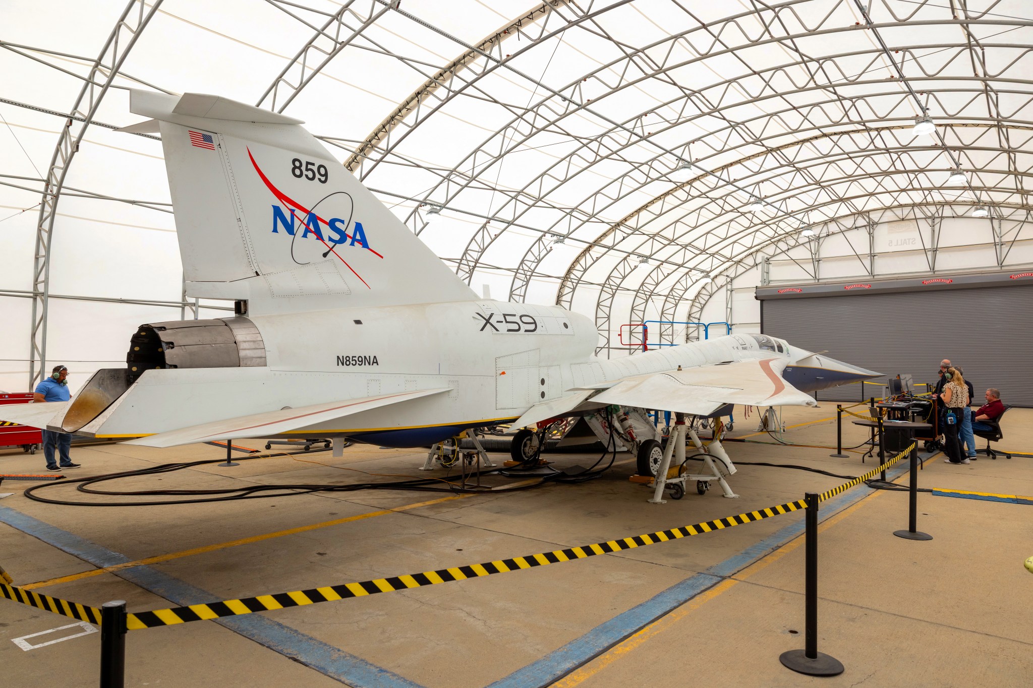 A white NASA jet airplane sits inside a fabric-covered-roof hangar with several people working at a nearby table with computers.